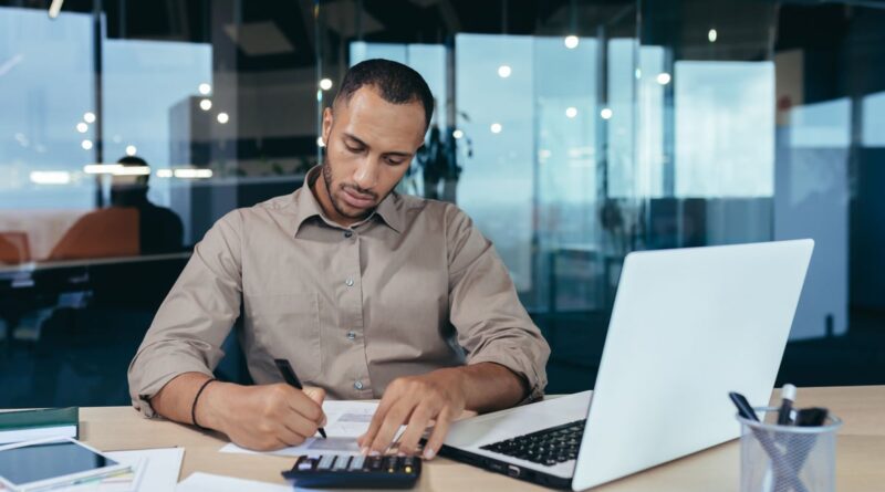 Investor using a calculator while sitting in front of a laptop.